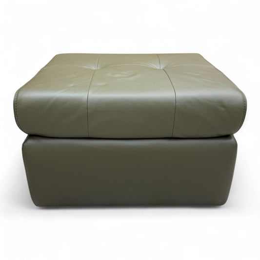 The 'Ava' khaki green leather footstool positioned in a living space, demonstrating its use as an ottoman or extra seating.