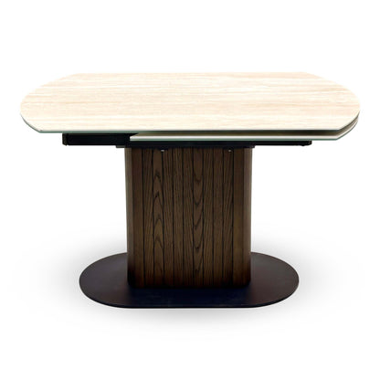 Front view of the Tiber travertine ceramic table closed, dark wood ribbed pedestal on an oval base
