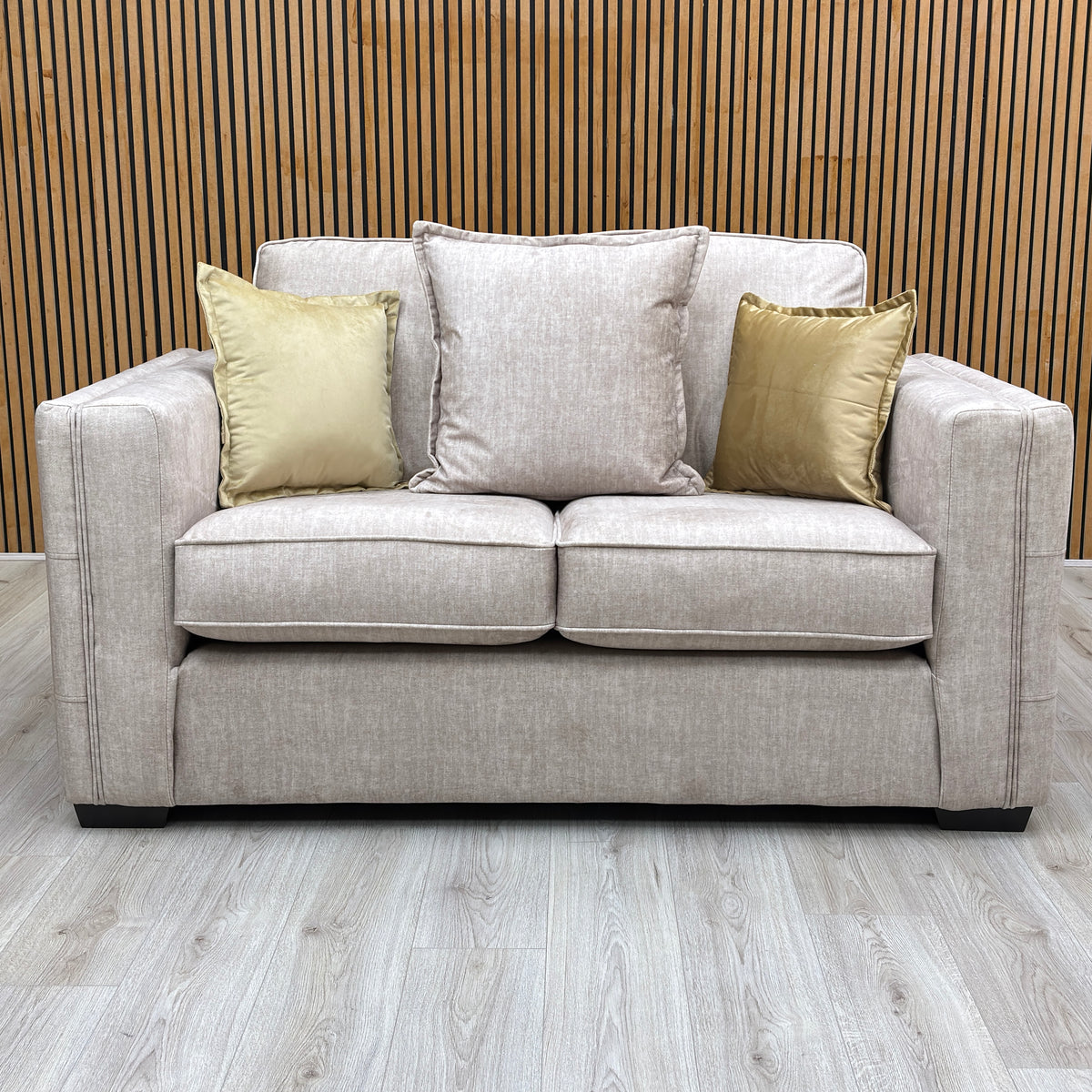 The Chelsea beige velvet 2 seater sofa from the set against a slatted wooden wall with gold cushions