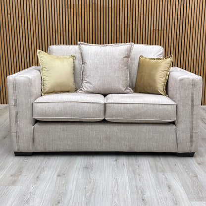 The Chelsea beige velvet 2 seater sofa from the set against a slatted wooden wall with gold cushions