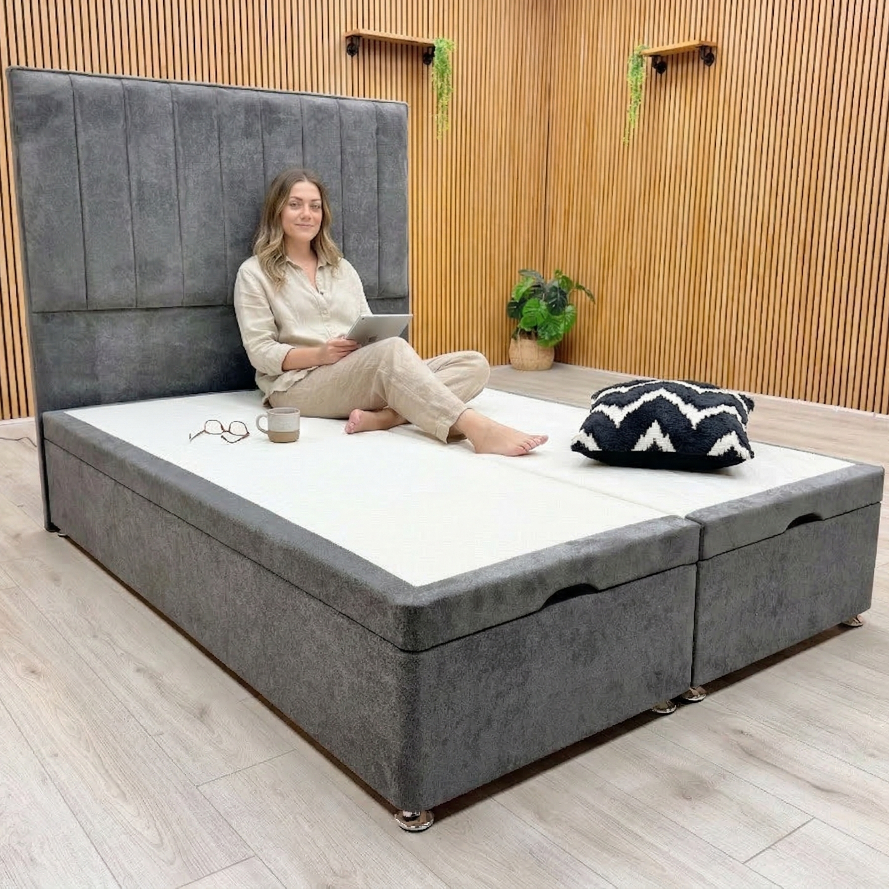 Woman sitting on a gray upholstered bed with storage drawers in a room with wooden walls and a plant.