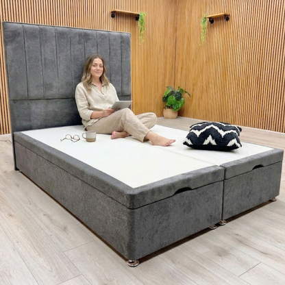 Woman sitting on a gray upholstered bed with storage drawers in a room with wooden walls and a plant.