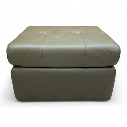 The 'Ava' khaki green leather footstool positioned in a living space, demonstrating its use as an ottoman or extra seating.