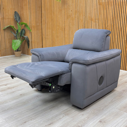 Lifestyle view of the Genius power recliner, highlighting the luxury Iron Grey velvet, built-in Bluetooth speakers, and dual-motor power headrest and recliner functions.
