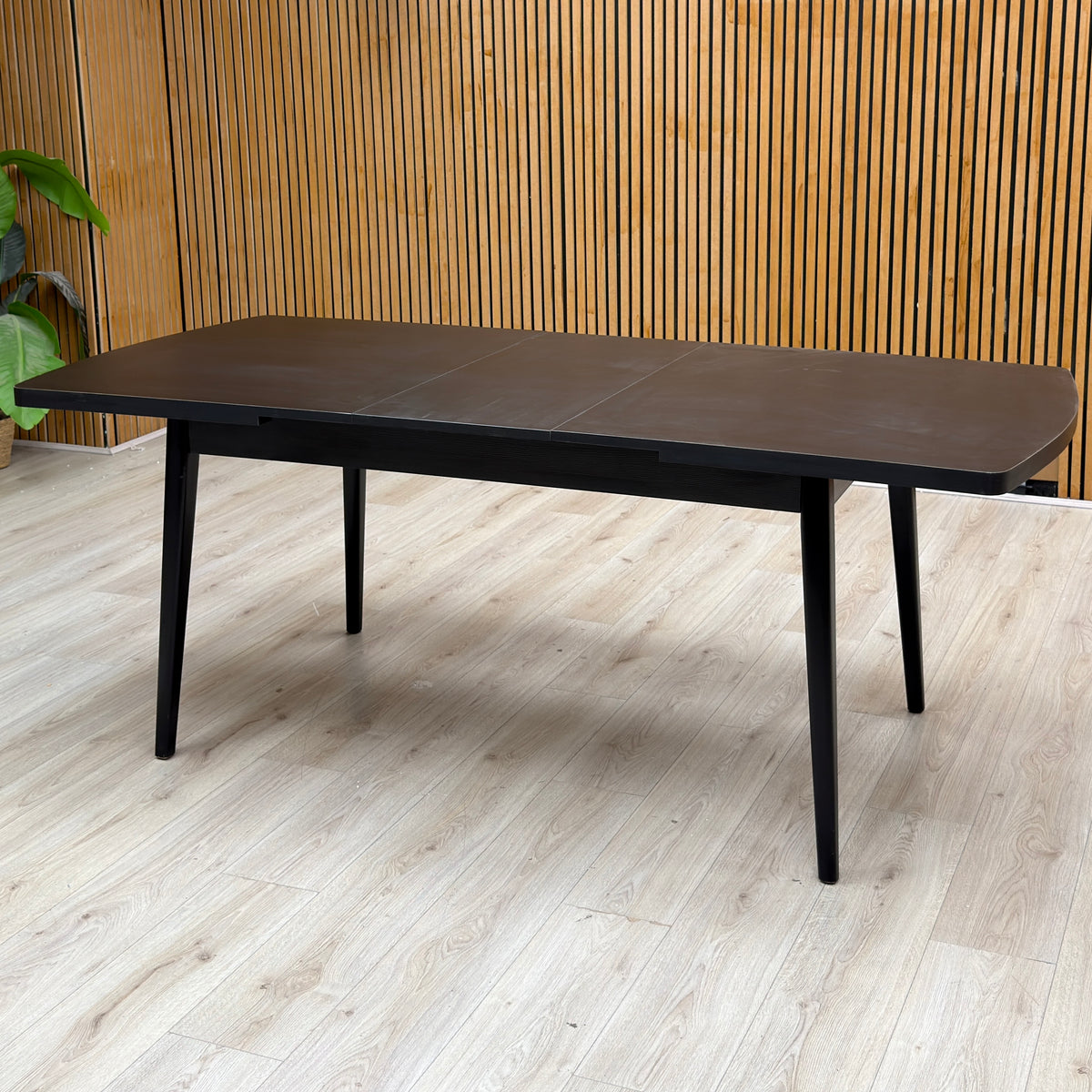 Magda black solid oak extending dining table extended, against a slatted wooden wall on a light oak floor