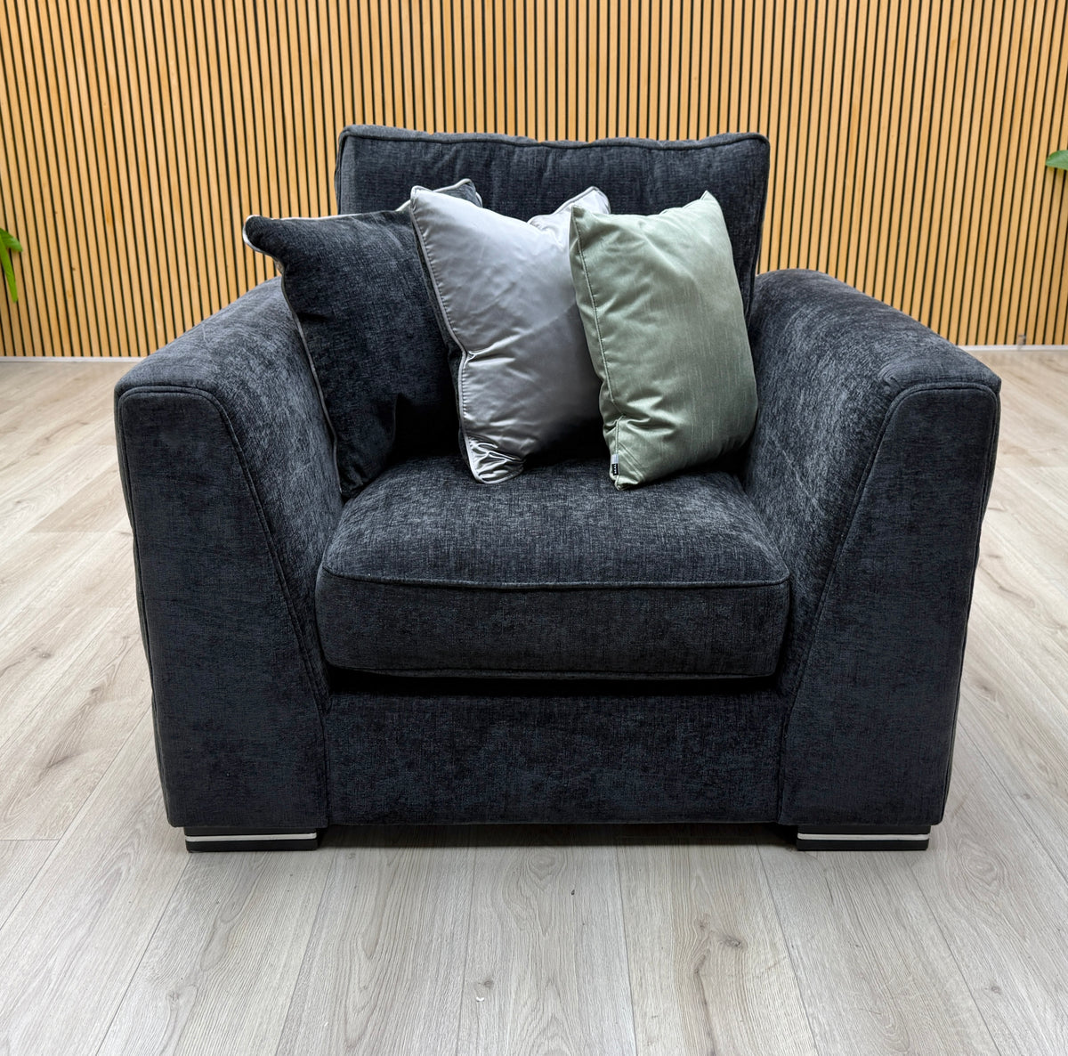 Front view of the armchair from the Bellissimo mirage black fabric set against a slatted wall with three cushions