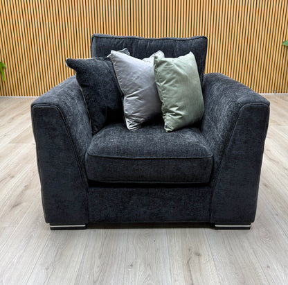 Front view of the armchair from the Bellissimo mirage black fabric set against a slatted wall with three cushions