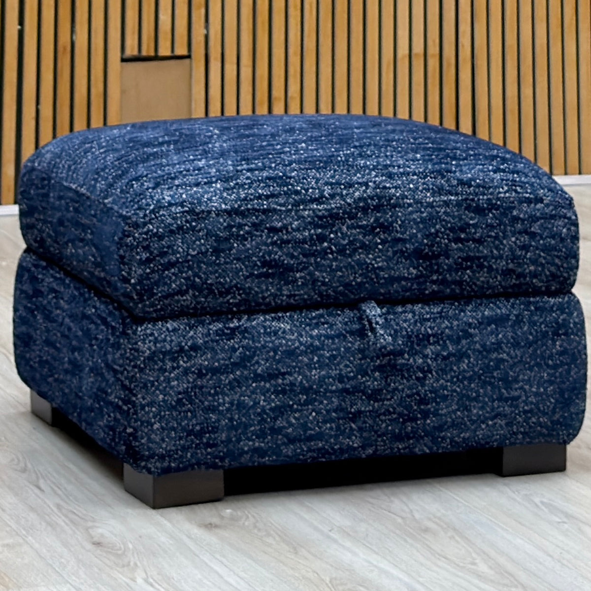 Nova dark denim blue woven fabric storage footstool on a light wood-effect floor by a slatted wall