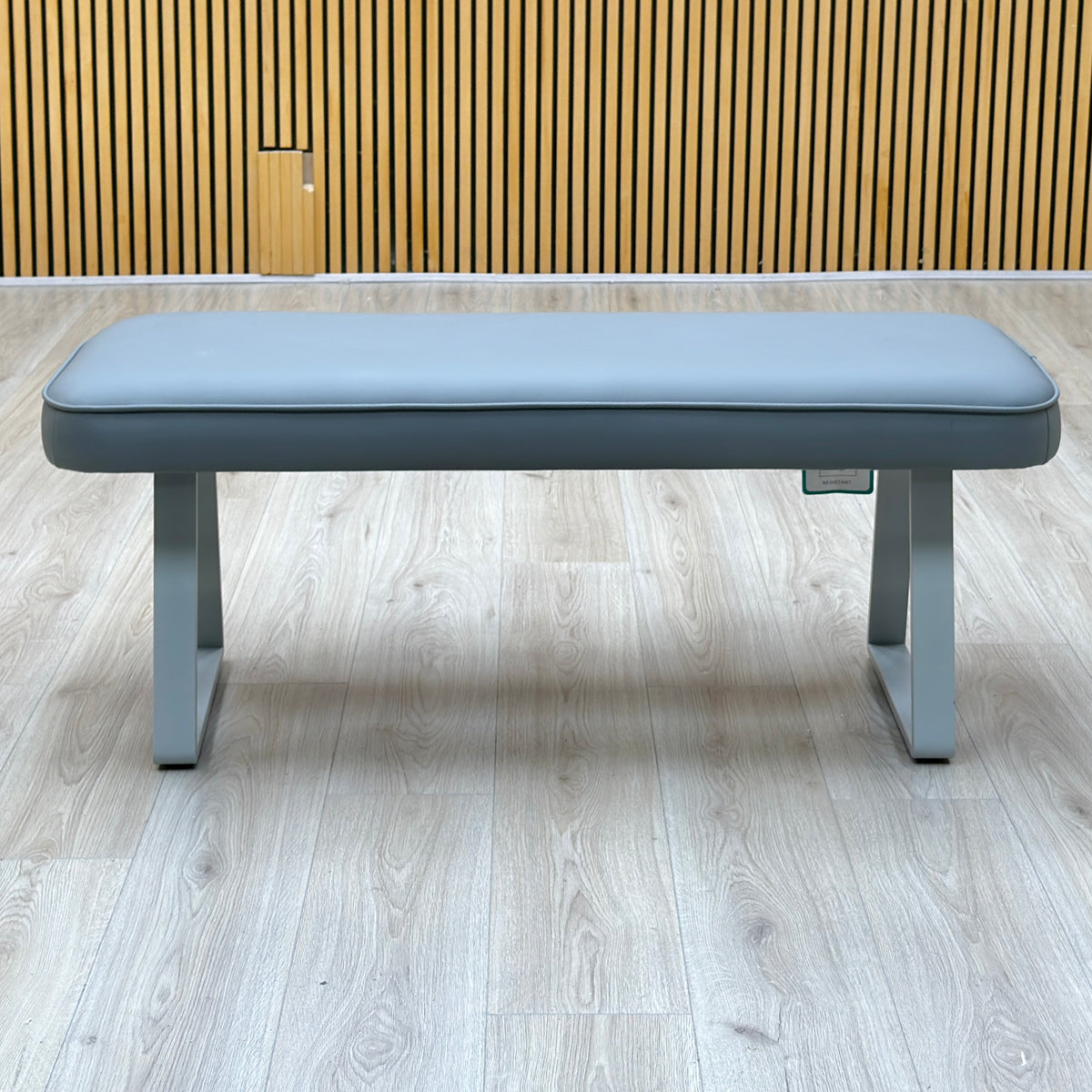 Front view of the Fumo light grey faux leather dining bench against a slatted wooden wall on a light oak floor