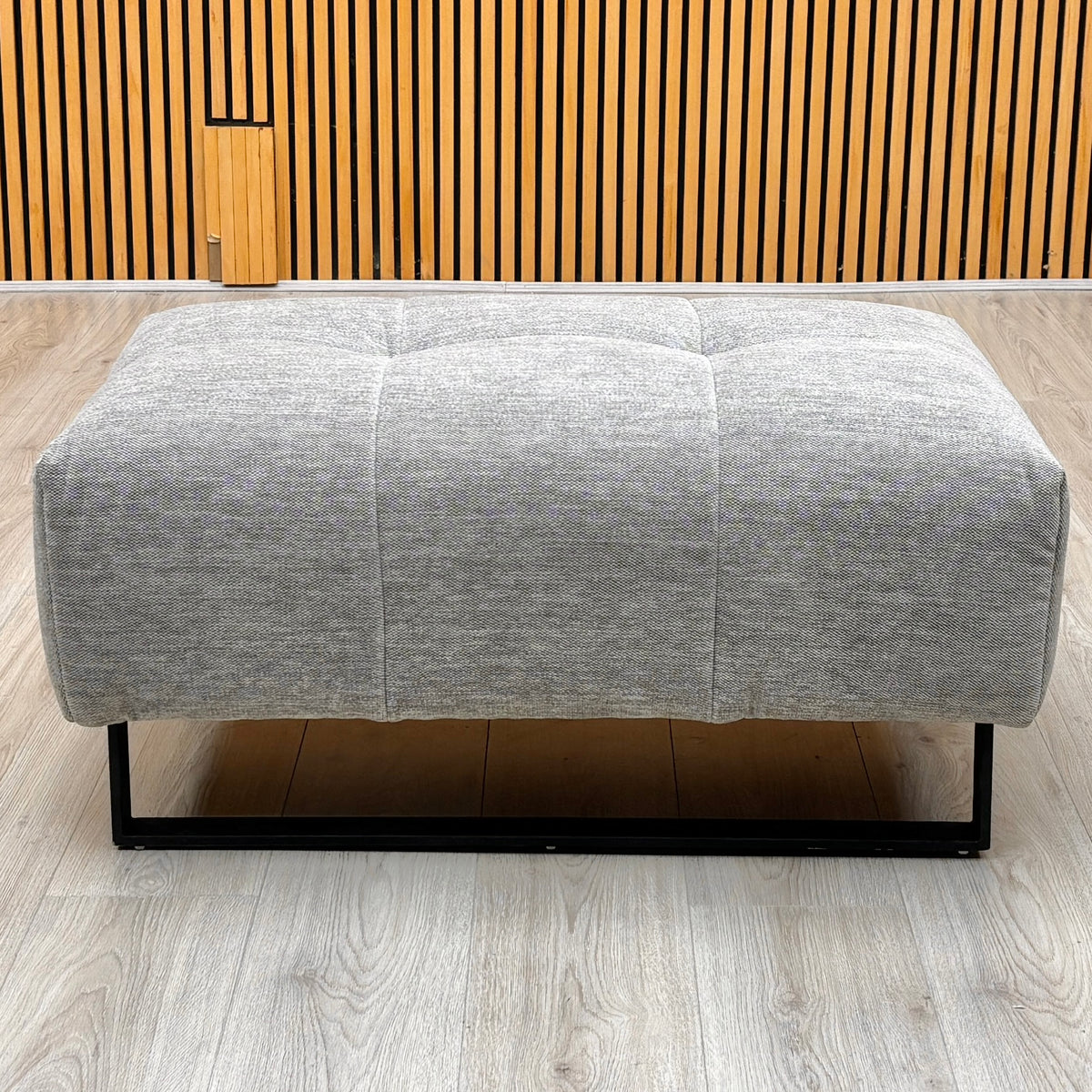 Shelby dove grey fabric footstool photographed against a slatted wooden wall on a light oak floor