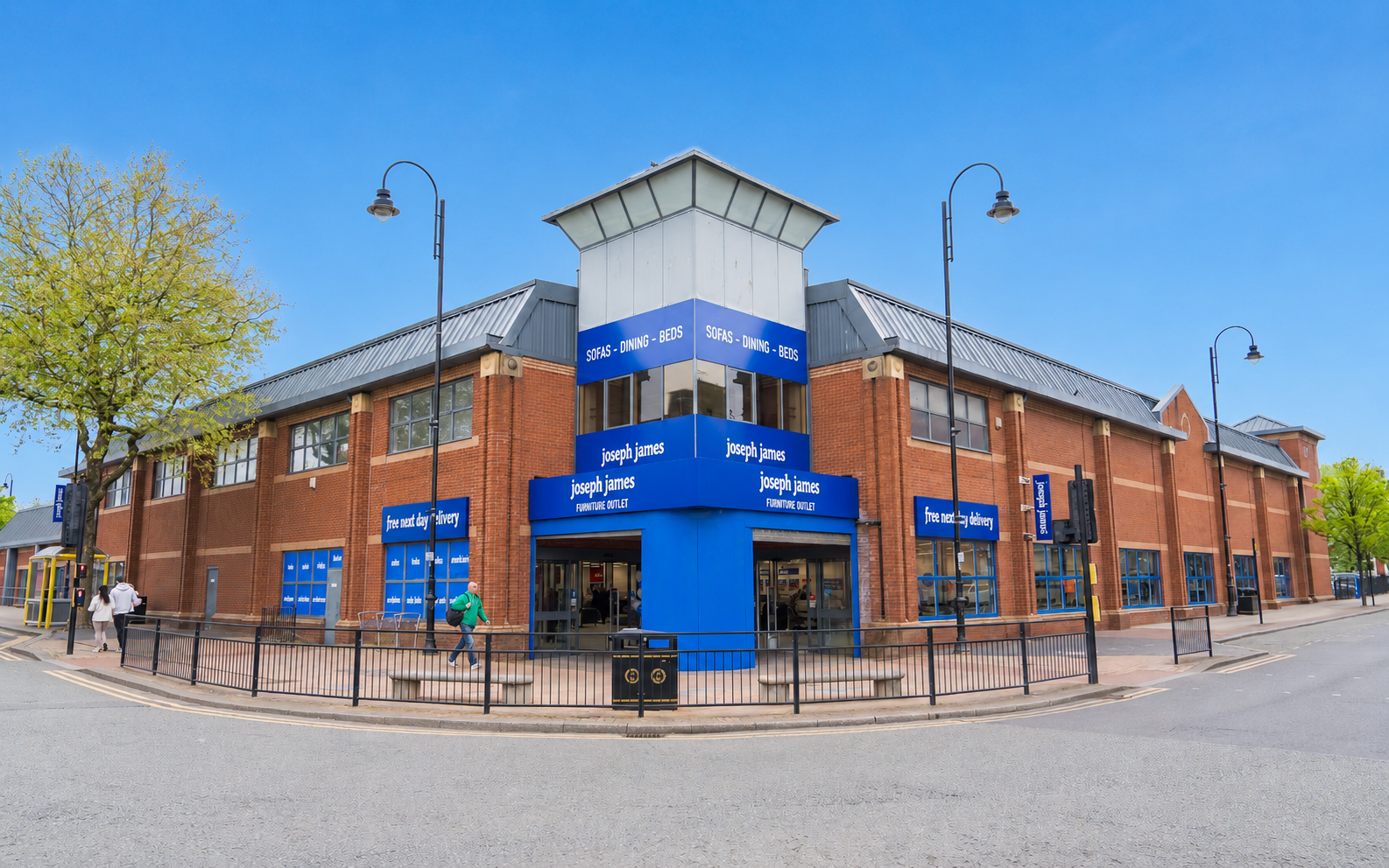 A corner brick retail building with large blue storefront signage reading Joseph Jones, set against a clear blue sky with trees and a paved street in the foreground.